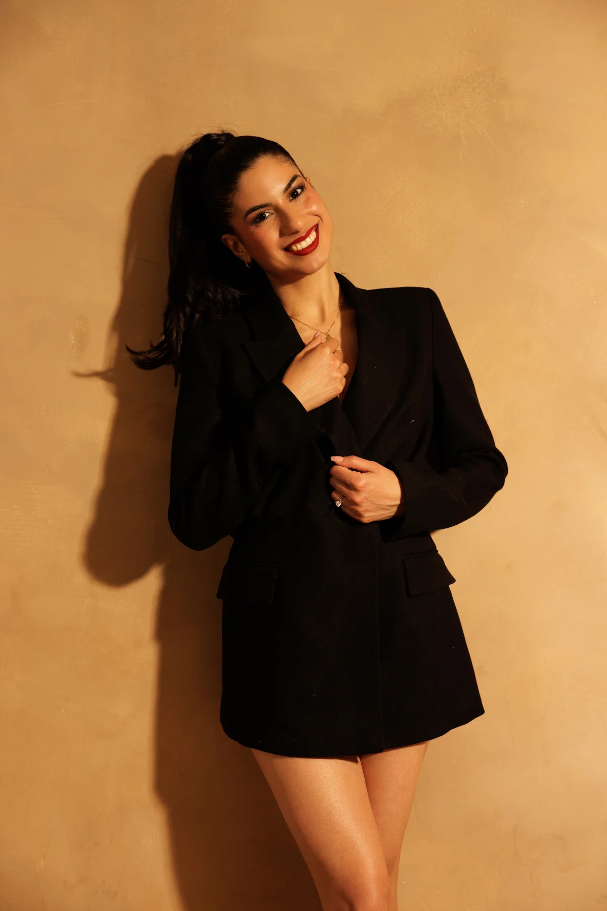 Claudia Cristina en blazer noir souriante contre un mur ocre