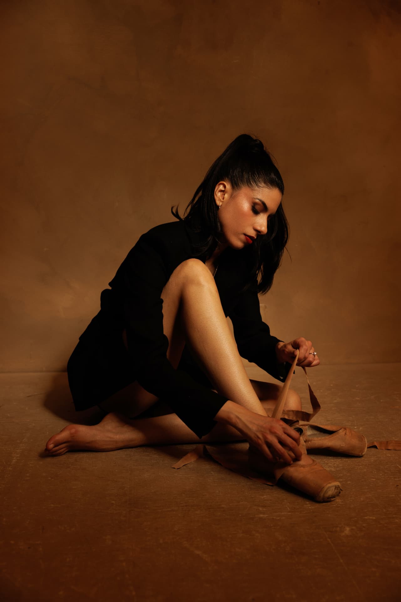 Claudia Cristina en blazer noir, assise, nouant ses pointes sur fond ocre